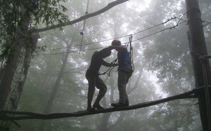 Two people wearing safety gear are secured by ropes as they navigate an obstacle of a high ropes course. The air around them appears foggy. 